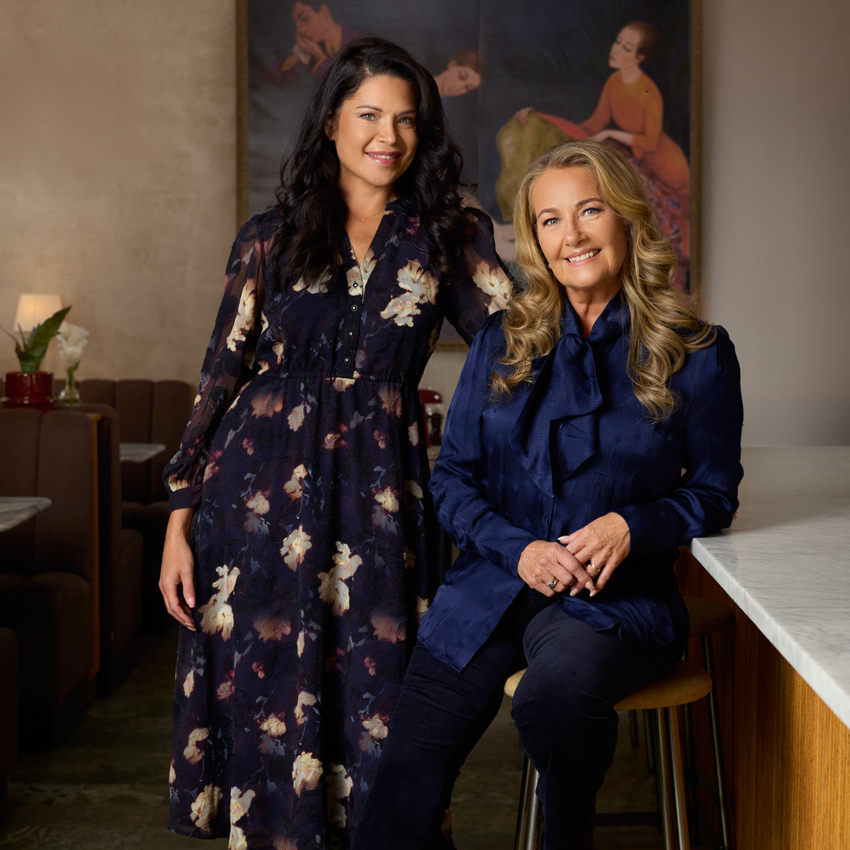 Yvonne Ryding har en mörkblå knytblus och matchande sammetsbyxor och står tillsammans med Jessica som har en blommig klänning i mörkblå, plommon och beige nyanser.