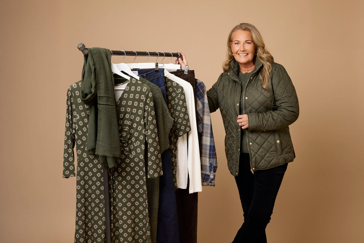 Yvonne Ryding med grön jacka och grön tröja och svarta manchesterbyxor står bredvid ett klädhänge för Naturlig stil
